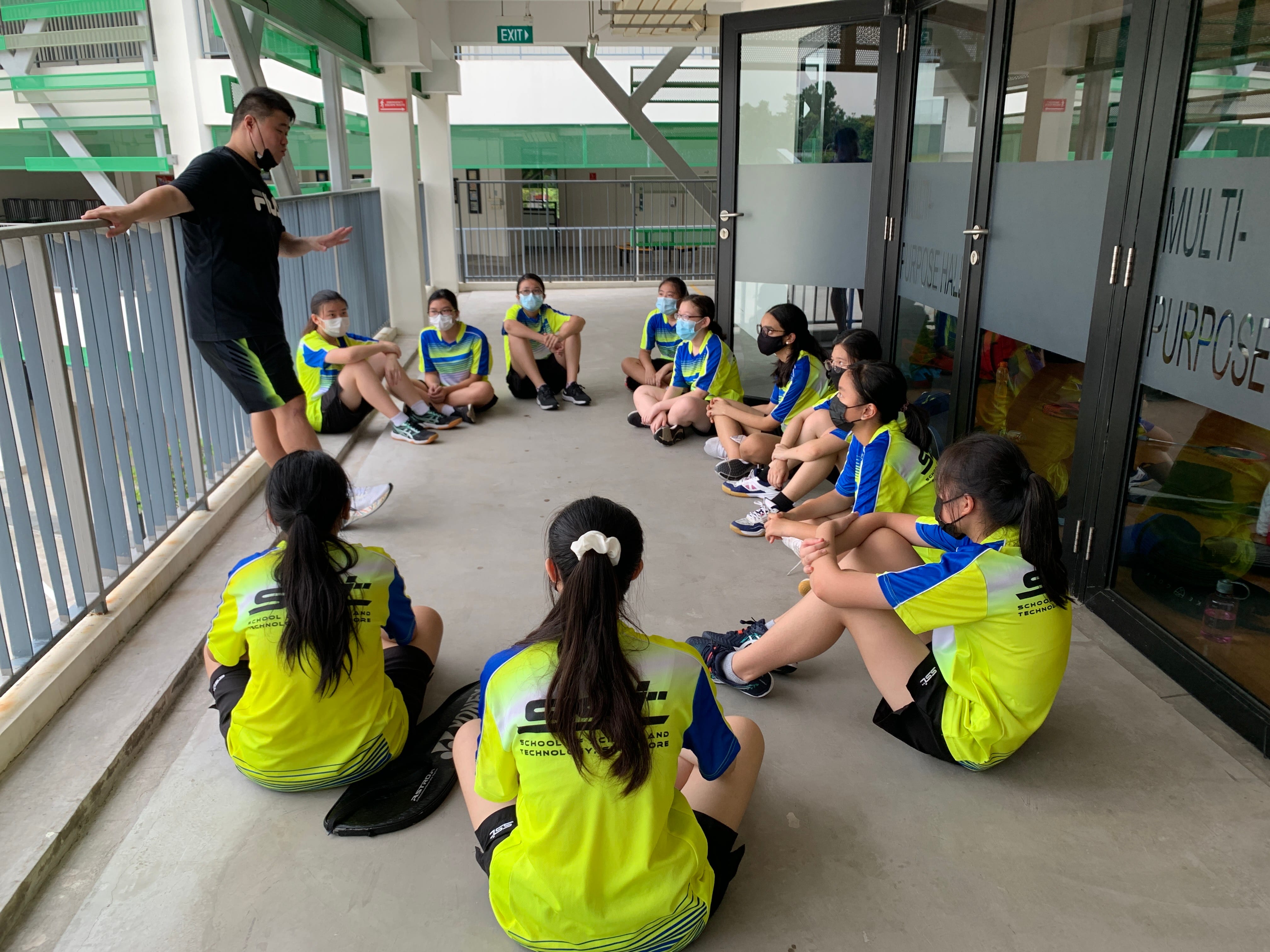 A coach in black addresses students in yellow-blue uniforms sitting in a semicircle on a corridor floor near a multi-purpose hall.